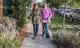 Two people walking side by side, one using a white cane, guided by a friend.