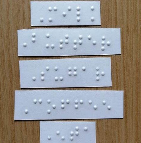 : Five white self-adhesive labels with embossed Braille text, arranged vertically on a wooden surface.