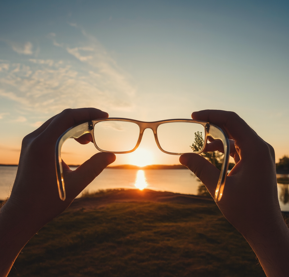 First-person view of someone holding eyeglasses toward a bright sunset, symbolizing new vision and perspective.